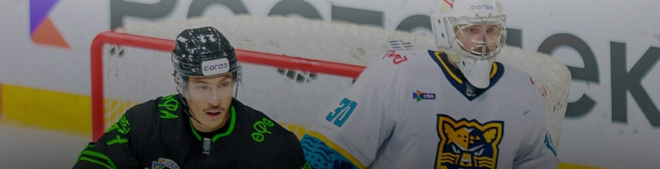 На изображении хоккеист в черной форме на льду опережает вратаря в серой форме на фоне ворот. Табло и реклама частично видны на заднем плане