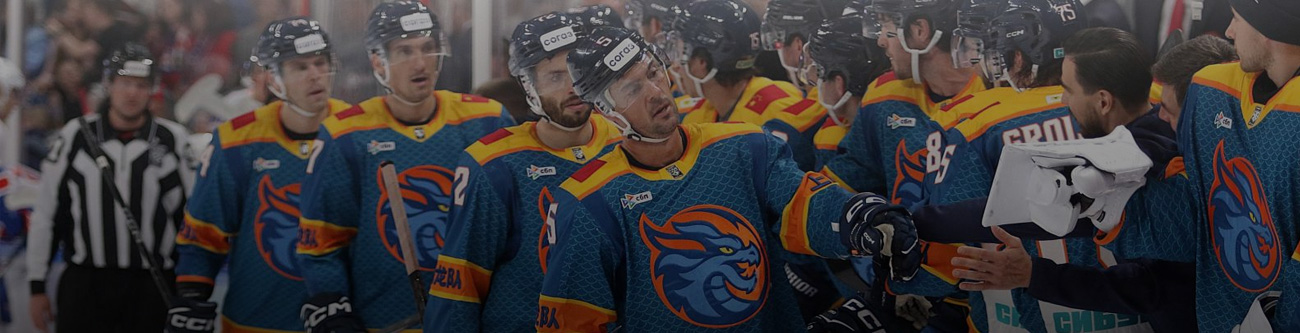 Хоккейная команда в синих формах с оранжевыми акцентами стоит на скамейке запасных. Игроки в шлемах разговаривают друг с другом. На фоне видны судья и другие участники матча