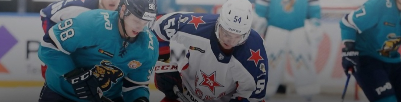 На изображении два хоккеиста в движении на ледовом поле. Один из них в светлой форме с красными звездами, другой в бирюзовой форме. Они активно борются за шайбу. Виден боковой борт хоккейной арены