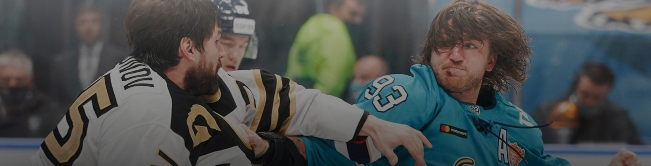 На изображении двое хоккеистов в защитной форме участвуют в борьбе. Игрок в темной форме схватился с игроком в светлой форме. На заднем плане виден еще один хоккеист в шлеме