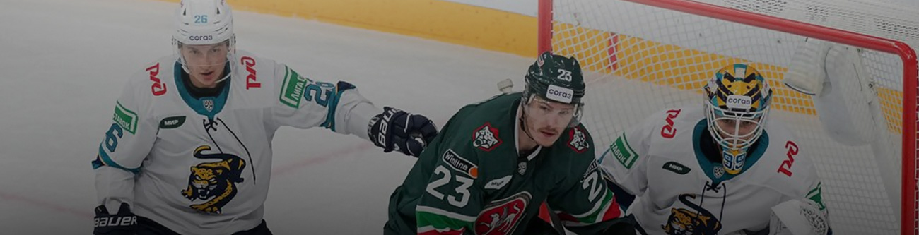 На изображении идет хоккейный матч. Игрок в зеленой форме и два игрока в белой форме, один из которых вратарь, находятся у ворот. Видна хоккейная атрибутика и сетка ворот